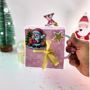 May include: A pink Christmas-themed mug with a Santa Claus sticker, a gold star, and a yellow ribbon. The mug has a white handle and is decorated with snowflake patterns. A small Christmas tree and a snow globe are in the background.