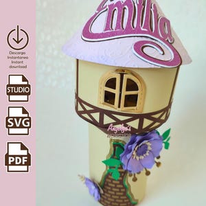 Puede incluir: Una torre decorativa de papel con un tejado lila claro que lleva el nombre "Emilia" en purpurina rosa. La torre tiene un cuerpo beige, un marco de ventana marrón y está adornada con flores de papel moradas y un diseño de cesta.
