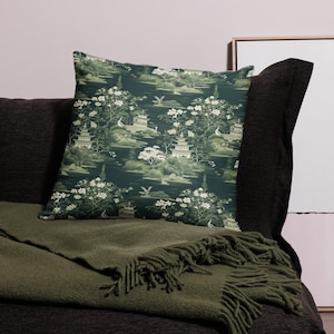 May include: Decorative throw pillow with a dark teal background featuring a repeating pattern of pagodas, trees, and birds in shades of green and white. The pillow is placed on a dark gray sofa with a green blanket.
