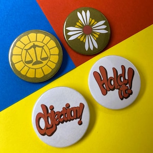 Op de afbeelding: Drie ronde buttons. Eén is geel met een zwart icoon van een weegschaal. Een andere is olijfgroen met een wit en geel bloemmotief. De overige twee zijn wit met rode tekst die "Objection!" en "Hold it!" weergeeft.