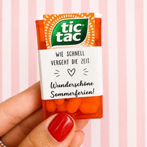 Könnte beinhalten: Ein rechteckiger, orangefarbener Tic Tac Behälter gefüllt mit orangefarbenen Dragees. Ein weißes Papieretikett ist um die Mitte gewickelt und zeigt den Text WIE SCHNELL VERGEHT DIE ZEIT, ein Herzsymbol und den Text Wunderschöne Sommerferien.