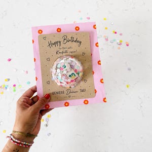 Könnte beinhalten: Eine rechteckige Geburtstagskarte mit rosa Rand und orangefarbenen Blumenmotiven. Die beigefarbene Mitte trägt den Text Happy Birthday, für Dich soll's Konfetti regnen! und GENIEBE Deinen TAG!. Eine transparente Kunststoffkuppel enthält buntes Konfetti.