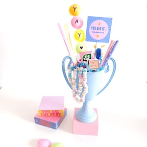 May include: A light blue trophy cup filled with colorful candy, pens, and decorative sticks. The cup holds a card reading YOU DID IT! and a pack of Tic Tac mints. A stack of two note pads reading YOU ARE THE BEST sits nearby with three round candies.