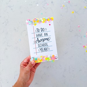 May include: A rectangular white card featuring blue horizontal lines and a red vertical margin line. The text reads: TO DO: HAVE AN Awesome SCHOOL YEAR! A small red heart is printed above the text. The top and bottom edges are decorated with colorful confetti.