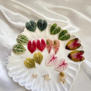 May include: A collection of leaf-shaped earrings in various colors, including green, red, pink, and yellow, displayed on a white scalloped plate. Several pairs of stud earrings and a pair of gold hoop earrings are also present.