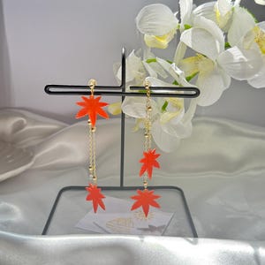 May include: A pair of dangle earrings with red maple leaf charms and gold chains. The earrings are displayed on a black stand.