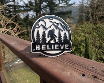 May include: A white and black metal sign with a silhouette of Bigfoot standing in a mountain range with trees. The sign says "BELIEVE" in black letters.