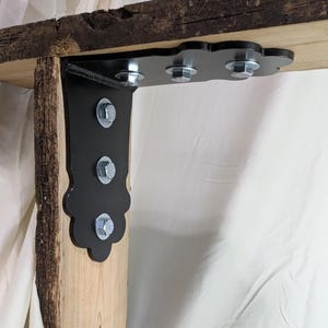 May include: A black metal bracket with decorative edges is bolted to a wooden beam. The bracket is attached with silver bolts. The wood has a natural, rough bark edge. The bracket is designed to support a shelf or other structure.