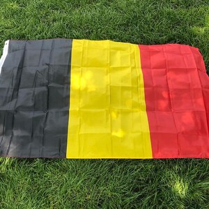 Könnte beinhalten: Eine rechteckige Flagge mit drei vertikalen Streifen gleicher Breite in Schwarz, Gelb und Rot. Die Flagge hat einen weißen Streifen an der linken Seite mit zwei Metallösen zur Befestigung. Der Stoff weist deutlich sichtbare Faltlinien auf.