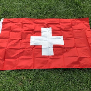 Puede incluir: Una bandera rectangular con un fondo rojo sólido y una cruz blanca centrada. La bandera incluye una franja vertical blanca en el lado izquierdo con dos ojales metálicos para su montaje, mostrando un diseño clásico con pliegues visibles.