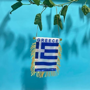 Könnte beinhalten: Eine kleine rechteckige Flagge von Griechenland mit blauen und weißen horizontalen Streifen und einem weißen Kreuz auf blauem Grund. Das Wort GREECE ist oben in Blau aufgedruckt. Die Flagge hat goldene Fransen und eine goldene Kordel.