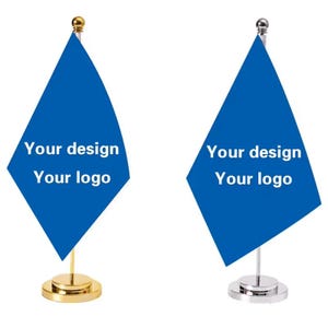 Op de afbeelding: Twee blauwe driehoekige bureaustandaarden met de tekst Your design Your logo in het wit gedrukt. De ene vlag heeft een goudkleurige metalen mast en ronde voet, de andere heeft een zilverkleurige metalen mast en ronde voet.