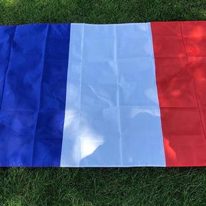 May include: A rectangular flag of France featuring three vertical stripes of equal width in blue, white, and red. The flag includes a white header strip along the left side with two metal grommets for mounting, displaying a smooth fabric texture.