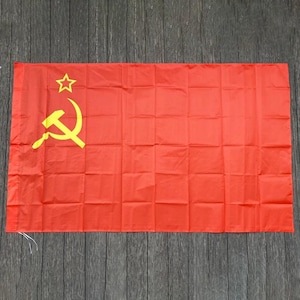 Può includere: Una bandiera rettangolare con uno sfondo rosso a tinta unita e un simbolo giallo di falce e martello posizionato nell'angolo in alto a sinistra, accompagnato da una stella gialla a cinque punte situata sopra l'emblema.