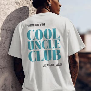 Könnte beinhalten: Weißes T-Shirt mit dem türkisfarbenen Text "PROUD MEMBER OF THE COOL UNCLE CLUB". Darunter steht "LIKE A DAD BUT COOLER". Die Person trägt eine dunkelblaue Kappe.
