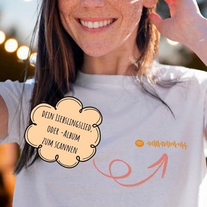 Op de afbeelding: Een wit T-shirt met een roze spraakballon die "DEIN LIEBLINGSLIED, ODER -ALBUM ZUM SCANNEN" zegt en een roze pijl die naar een barcode wijst.
