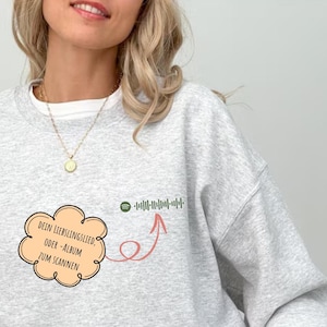 May include: Gray sweatshirt with a white cloud and red arrow pointing to a Spotify barcode. The text on the cloud reads "DEIN LIEBLINGSLIED, ODER -ALBUM ZUM SCANNEN".