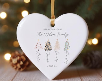 May include: White heart-shaped ceramic Christmas ornament with a gold ribbon. It features a stylized Christmas tree design with the text "Merry Christmas The Wilson Family" and the names "Aurelia, Joseph, Everly, Pristley" and the year "2024".