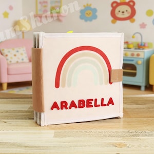 May include: A square fabric quiet book with a cream-colored cover featuring a red, tan, and light blue rainbow graphic. The name ARABELLA is printed in red capital letters below the rainbow. The book has a tan side spine and a small side tab closure.