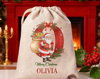 May include: A beige burlap Christmas gift bag with a drawstring closure. The bag features a circular wreath design with a Santa Claus illustration and the text "Merry Christmas Olivia". The wreath is red with green accents.