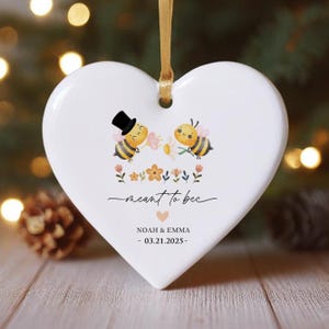 Può includere: Ornamento bianco a forma di cuore con un nastro dorato. L'ornamento presenta due api dei cartoni animati, elementi floreali e la scritta "meant to bee". Sotto, i nomi "Noah & Emma" e la data "03.21.2025."