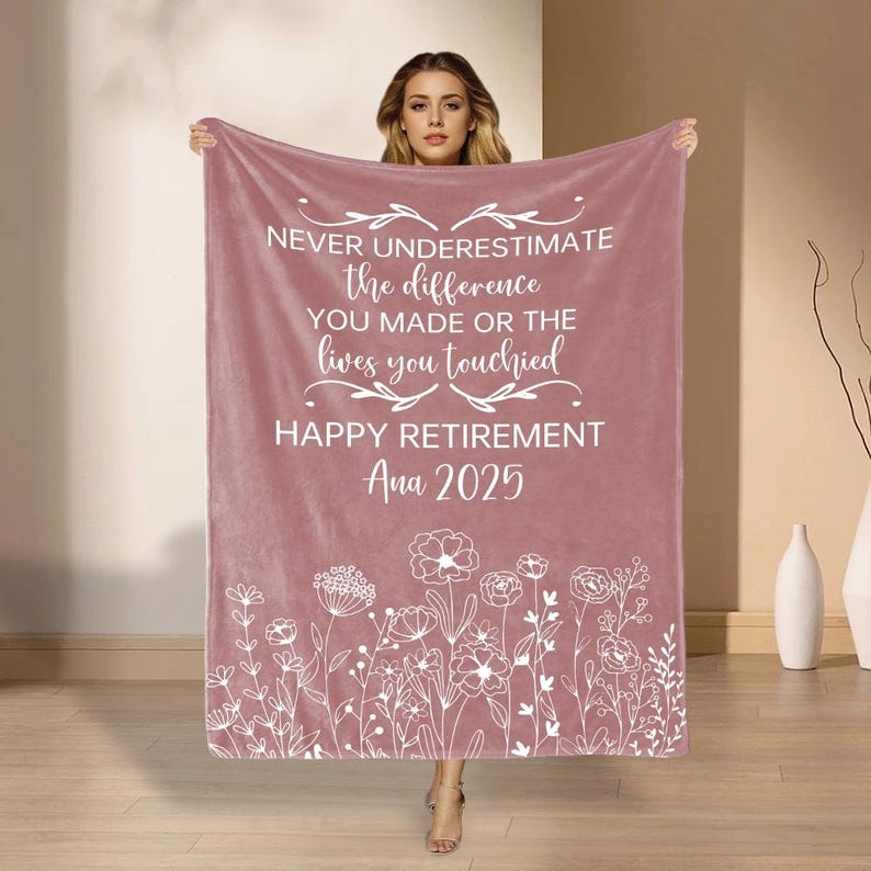 May include: A dusky pink blanket with white text that reads "NEVER UNDERESTIMATE the difference YOU MADE OR THE lives you touched HAPPY RETIREMENT Ana 2025." The bottom edge features a white floral design.