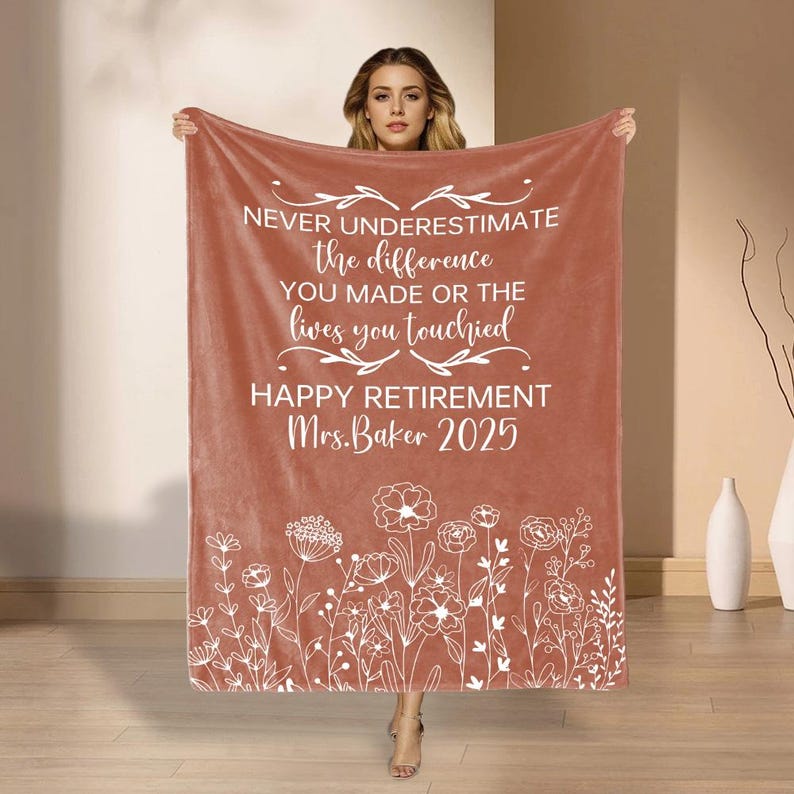 May include: A terracotta blanket with white text: "NEVER UNDERESTIMATE the difference YOU MADE OR THE lives you touched HAPPY RETIREMENT Mrs. Baker 2025." A white floral pattern decorates the bottom edge of the blanket.