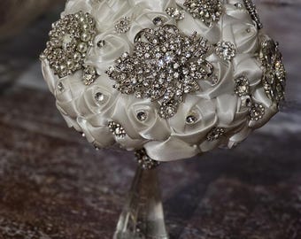 May include: A white satin bridal bouquet with rhinestone and pearl embellishments. The bouquet features a variety of silver brooches and sparkling accents. The handle is clear, giving the illusion that the bouquet is floating.