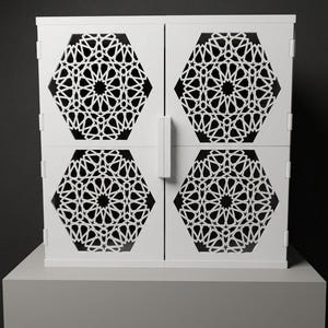 May include: A white cabinet featuring two doors with intricate, laser-cut geometric patterns in a hexagonal shape. The design consists of repeating star and polygon motifs, creating a lattice effect that reveals a dark interior through the cutouts.