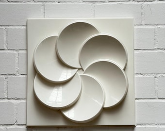 May include: A white, geometric wall art piece featuring a square base with six overlapping, semi-circular shapes arranged in a flower-like pattern. The artwork is mounted on a white brick wall.