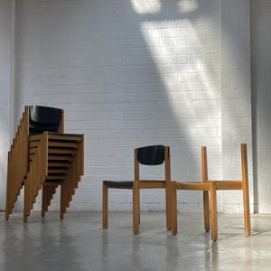 May include: A stack of several wooden chairs with black seats and backrests, positioned next to two individual chairs of the same design. The chairs feature light-toned wooden frames with straight legs and simple, functional construction.