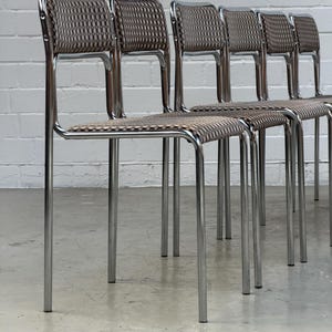 Op de afbeelding: Een set van zes chromen stoelen met geweven bruine en beige zittingen en rugleuningen. De stoelen hebben een minimalistisch ontwerp met slanke, rechte poten en een gebogen rugleuning. De stoelen zijn in een rij gerangschikt.
