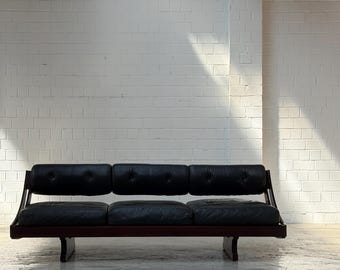 May include: A mid-century modern three-seat sofa with black leather cushions and a dark wood frame. The sofa has a low profile and a minimalist design, perfect for a living room or lounge area.