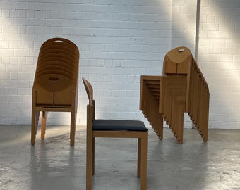 May include: Wooden chairs with a unique design. One chair is standing, while others are stacked. The chairs have a light brown finish and a dark seat. The background is a white brick wall and a gray floor.