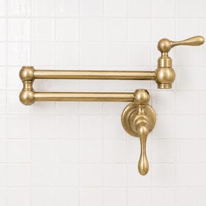May include: A wall-mounted pot filler faucet with a brushed brass finish. The fixture features a double-jointed, articulating swing arm design with two handles for water control, connected to a circular base plate that mounts directly to the wall.