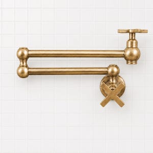 May include: A wall-mounted pot filler faucet with a brushed brass finish. The fixture features a double-jointed articulating arm, a cross-handle control valve at the base, and a secondary cross-handle at the spout end for water flow regulation.