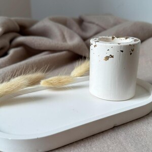 May include: White oval tray with a white cylindrical candle holder with gold flecks. The tray and holder are on a light brown surface.