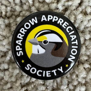 May include: A black and yellow button with the text "Sparrow Appreciation Society" and a cartoon sparrow with a yellow head and brown chest.