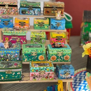 K&ouml;nnte beinhalten: Eine Sammlung farbenfroher, handbemalter Holzkisten mit aufwendigen Blumen- und Naturmotiven. Die Kisten werden auf einem mehrstufigen Holzregal pr&auml;sentiert und zeigen eine Vielzahl von Gr&ouml;&szlig;en und leuchtenden Farben, darunter Blau, Gr&uuml;n, Rosa und Gelb.