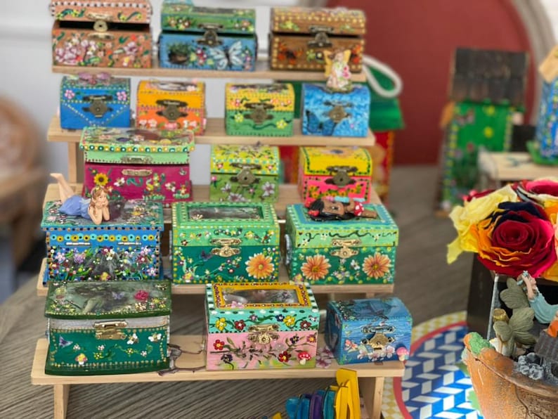 K&ouml;nnte beinhalten: Eine Sammlung farbenfroher, handbemalter Holzkisten mit aufwendigen Blumen- und Naturmotiven. Die Kisten werden auf einem mehrstufigen Holzregal pr&auml;sentiert und zeigen eine Vielzahl von Gr&ouml;&szlig;en und leuchtenden Farben, darunter Blau, Gr&uuml;n, Rosa und Gelb.