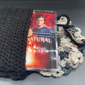 Peut inclure: Un sac en crochet noir avec un DVD de Supernatural à l'intérieur. La jaquette du DVD présente un homme aux cheveux foncés portant une chemise noire. Le sac est orné d'un pendentif en forme de croix argentée et d'une clé.