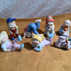 May include: A collection of six small porcelain figurines depicting children in various poses. The figures feature painted facial expressions, colourful clothing, and accessories including a baseball glove, a teddy bear, and a small rattle toy.