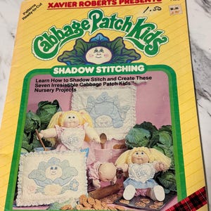 May include: A vintage Cabbage Patch Kids shadow stitching pattern book. The cover is yellow with green and red accents, featuring the Cabbage Patch Kids logo and illustrations of dolls and nursery projects. The book includes the text "Xavier Roberts Presents".