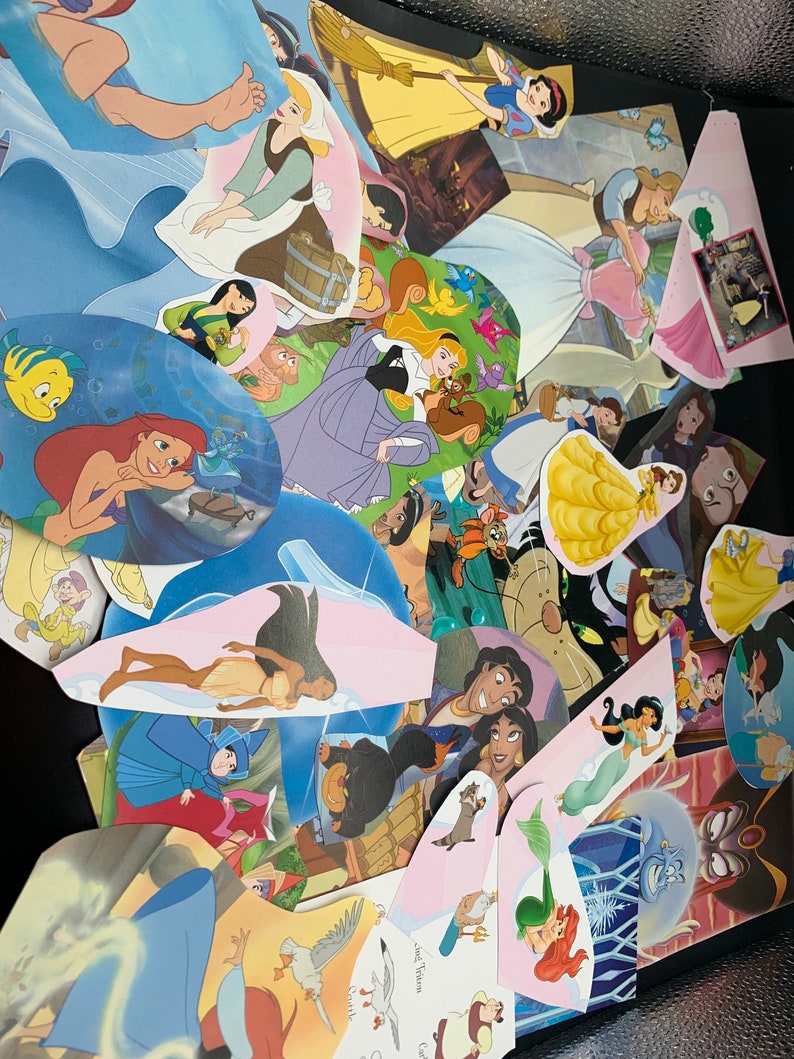 May include: A collection of colorful Disney princess character cutouts, including Ariel, Belle, Cinderella, Snow White, and Pocahontas. The cutouts are arranged in a random pattern on a dark surface.