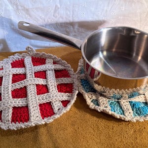 May include: A stainless steel saucepan rests on a round, blue and white crocheted trivet designed to look like a lattice-top pie. Beside it lies a matching round, red and white crocheted trivet, also featuring a lattice-top pie design.