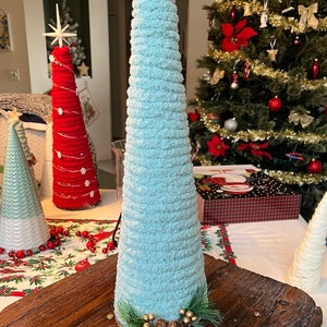 May include: A light blue, fuzzy, cone-shaped Christmas tree with a gold star on top. The tree is decorated with pine sprigs and gold ornaments. The tree is sitting on a wooden table.