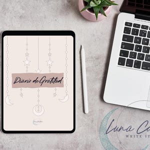 Puede incluir: Una tableta digital muestra una portada de diario con el texto "Diario de Gratitud". El diseño incluye estrellas colgantes, lunas y una luna creciente. Un lápiz óptico blanco y un portátil son visibles.