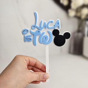 May include: A blue and white cake topper with the name "Luca" and the number "2" in a Disney-inspired font. A black Mickey Mouse head silhouette is on the right. The topper is held by a white stick.
