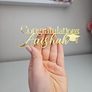 May include: Gold glitter acrylic cake topper with the text "Congratulations Aishal" and a graduation cap.
