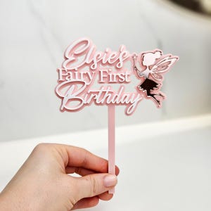 May include: A pink and white cake topper with the text "Elsie's Fairy First Birthday" and a fairy illustration. The topper is held by a hand against a white background. The fairy has a wand and wings.